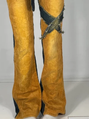 Unika tvåfärgade bootcut jeans - Säljer ett par riktigt unika bootcut jeans från Samurai Jeans med cool tvåfärgad design i mörkblått och senapsgult. Byxorna har råa detaljer, patchade fickor och ett avtagbart bälte i denim. Materialet är en mix av jeans och bomull, med slitna detaljer och korsade sömmar på låret.
