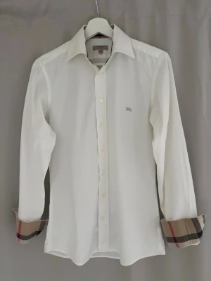 Vit Burberry skjorta slim fit XS - Snygg vit skjorta från Burberry i slim fit med klassisk krage och långa ärmar. Ikoniskt rutigt mönster på insidan av krage och manschetter. Skjortan har knappar framtill och är gjord i mjuk bomull. Perfekt för dig som gillar stilrena detaljer och designerplagg.