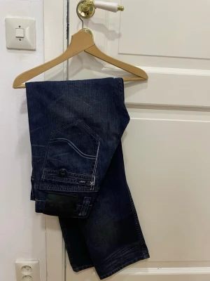 Vintage örkblå jeans från Lee - Snygga Vintage mörkblå jeans från Lee med klassisk femficksdesign och blåa detaljer på bakfickan. Jeansen har en rak passform, lite utsvängda, och är tillverkade i slitstarkt denimmaterial. Perfekta för dig som gillar en tidlös och clean look.
