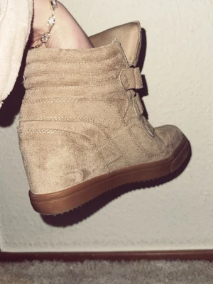 Beige wedge sneakers med kardborre - Säljer ett par beige wedge sneakers med tre breda kardborreband och platt brun sula. Skorna är tillverkade i mocka och har en rund tå samt en inbyggd klack för extra höjd. Perfekt för dig som vill ha en cool och bekväm stil.