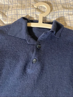 Mörkblå stickad pikétröja - Säljer en mörkblå stickad pikétröja med klassisk krage och två knappar framtill. Tröjan är av 100% nyull. 