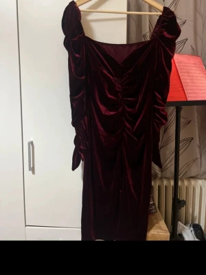 Vinröd sammetsklänning med volang - Vinröd sammetsklänning med snygga volangdetaljer längs axlarna och ärmarna. Klänningen har en tight passform och rynkade detaljer framtill som ger en cool look. Perfekt för dig som vill sticka ut med en elegant men edgy stil.