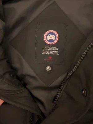 Svart pufferjacka från Canada Goose - Svart pufferjacka från Canada Goose med huva och klassisk patch på ärmen. Jackan har quiltad design, dragkedja och knappar framtill. Perfekt för kalla dagar och har en riktigt schysst look som funkar till streetwear. Materialet är polyester och dun.