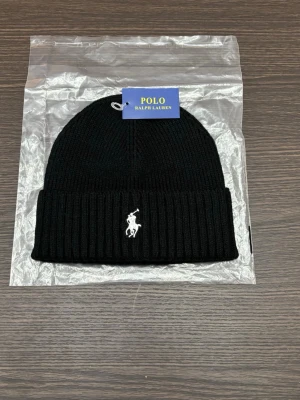 Svart stickad mössa från Polo Ralph Lauren - Snygg svart stickad mössa från Polo Ralph Lauren med klassisk ribbad design och vit broderad logga framtill. Perfekt för att hålla värmen och samtidigt se stilren ut. Passar dig som gillar enkel och tidlös stil.