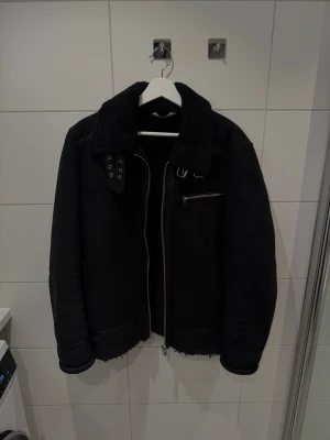 Zara jacka med pälskrage - strl L - Säljer en snygg svart jacka från Zara med skön pälskrage och detaljer i skinn. Jackan har en dragkedja framtill, en ficka med dragkedja på bröstet och spänndetaljer vid halsen. Materialet är mjukt och värmande (mockaimitation), perfekt för kyliga dagar.