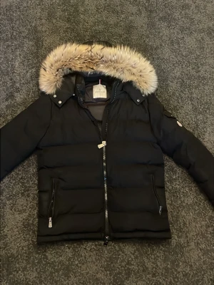 Svart Moncler pufferjacka med päls - Svart pufferjacka från Moncler med avtagbar huva och fluffig päls runt kanten. Jackan har två fickor med dragkedja, quiltad design och Moncler-logga på ärmen. Perfekt för kalla dagar och har en clean, modern look.