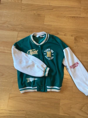 Stranger Things bomberjacka Hawkins High - Grön och vit bomberjacka med tryck från Hawkins High School och Tigers, samt Stranger Things-logga på ena ärmen. Jackan har ribbade muddar, knappar framtill och detaljer i gult och vitt. Perfekt för dig som gillar retrostil och popkultur.
