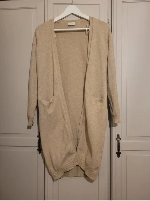 Beige lång kofta från Vila XS - Lång, beige kofta från Vila i storlek XS. Koftan har öppen front, två fickor fram och ribbade muddar vid ärmslut och nederkant. Perfekt för lager på lager och chill vibes under kyligare dagar.