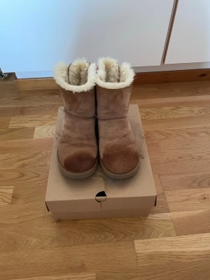 UGG Bailey Bow Chestnut🎀 - Säljer ett par klassiska UGG boots i beige mocka med fluffigt fuskpälsfoder och brun rosett baktill. Skorna har rund tå, platt sula och är perfekta för kalla dagar. UGG-logga på hälen och grov yttersula för extra grepp.