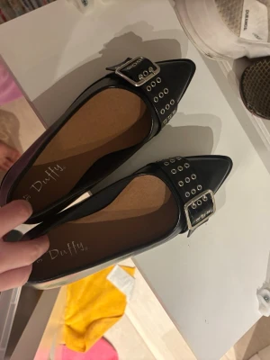 Svarta ballerinaskor med spänne från Duffy - Helt nyskick! Snygga svarta ballerinaskor från Duffy med spetsig tå och coola silvriga detaljer. Skorna har ett brett band med metallhål och ett stort spänne framtill som ger en edgy vibe. 