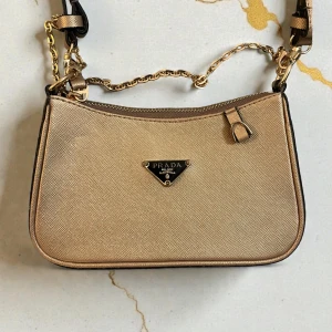 Guldig axelväska från Prda - Säljer en snygg guldig axelväska från Prda med klassisk trekantslogga framtill. Väskan har guldfärgade kedjedetaljer och justerbar axelrem. Tillverkad i strukturerat skinn som ger en lyxig känsla. Perfekt storlek för det viktigaste och en riktig statement piece.