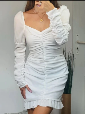 Supersnygg lyxig vit klänning ny med prislapp - Supersöt vit kortklänning med långärmade puffärmar och snygg omlottdesign. Klänningen har rynkade detaljer längs hela framsidan och en volangkant längst ner. Materialet är lätt och känns lyxigt, perfekt för dig som vill sticka ut med en fräsch look.