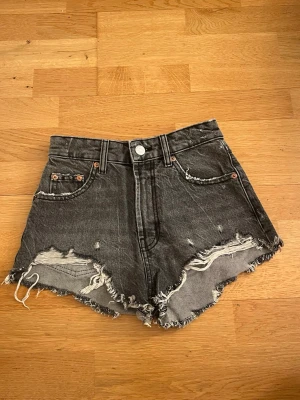 Grå jeansshorts från Zara - Säljer ett par grå högmidjade jeansshorts från Zara i storlek 32. De har råa, fransiga kanter och slitna detaljer framtill för en cool vibe. Klassisk knapp och dragkedja. Perfekta för varma dagar och chill outfits.