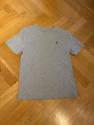 Grå t-shirt från Polo Ralph Lauren - Snygg grå t-shirt från Polo Ralph Lauren med klassisk rund hals och korta ärmar. Diskret Polo-logga broderad på bröstet. Tillverkad i mjuk bomull som känns skön mot huden. Perfekt för en chill och stilren look.