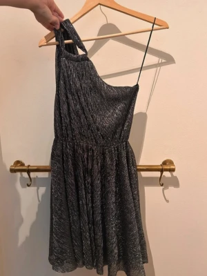 Glittrig svart oneshoulder klänning Maje - Supersnygg svart kortklänning från Maje med glittrigt tyg och oneshoulder-design. 