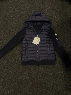 Mörkblå Moncler pufferjacka med huva - Säljer en mörkblå pufferjacka från Moncler med stickade ärmar och huva. Jackan har tvåvägsdragkedja, ribbade muddar och fickor med tryckknappar. Klassisk Moncler-logga på ärmen och coola detaljer på insidan. Perfekt för dig som vill ha både stil och funktion.😊