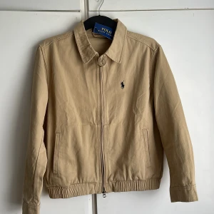 Beige jacka från Polo Ralph Lauren - Stilren beige jacka från Polo Ralph Lauren med klassisk krage, dragkedja framtill och broderad logga på bröstet. Jackan har elastisk nederkant och raka fickor. Perfekt för dig som gillar en clean och tidlös look.