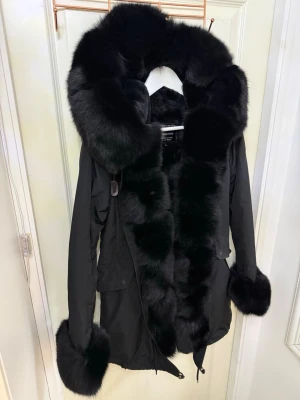 Svart pälsparkas  - Säljer en svart parkajacka från moomookong med fluffiga pälsdetaljer på huva, krage och ärmslut. Jackan har snörning nedtill och en loose passform som ger en cool vibe. Perfekt för kalla dagar när du vill vara både varm och snygg. Köptes för 3100kr.