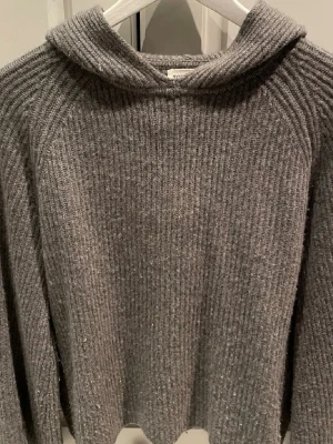 Grå hoodie i kashmir från Soft Goat - Mysig grå hoodie från Soft Goat i 100% kashmir. Tröjan har ribbad struktur, huva och långärmad design. Perfekt för dig som vill ha något både stilrent och superbekvämt. Passformen är normal och materialet är riktigt mjukt.