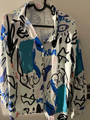 Vit skjorta med grafiskt mönster, strl L - Säljer en vit skjorta i storlek L med ett riktigt coolt grafiskt mönster i blått, svart, rosa och turkost. Skjortan har långa ärmar och knappar framtill. Materialet känns lätt och skönt, perfekt för dig som gillar att sticka ut med färg och konstnärliga prints.