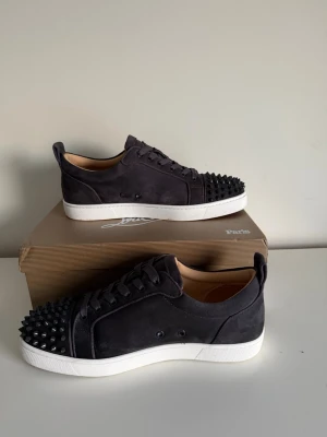 Gråa Christian Louboutin sneakers med nitar - Säljer ett par gråa sneakers från Christian Louboutin i mocka med coola svarta nitar på tån och klassisk röd sula. Skorna har snörning och vit platt sula. Kommer med originalkartong. Perfekta för dig som vill sticka ut med en edgy vibe.