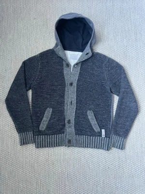 Grå cardigan jacka med huva från Replay - Säljer en mörkgrå stickad cardigan jacka från Replay med kontrasterande ljusgrå ribbade detaljer och huva i sweatshirtmaterial. Jackan har knappar framtill, två fickor och ribbade muddar. Perfekt för dig som gillar en chill och avslappnad stil. Den saknar en knapp i mitten. Den är XL på lappen men passar som L.