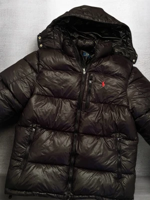 Svart pufferjacka Ralph lauren - Svart pufferjacka med huva från Polo Ralph Lauren. Jackan har en röd broderad logga på bröstet, flera dragkedjor och quiltad design. Materialet är glansigt syntet och dunfyllt, perfekt för kalla dagar. Snygg och praktisk med fickor och hög krage.