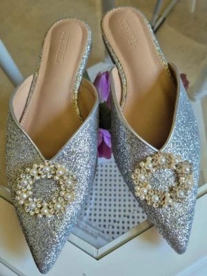 Glittriga silver ballerinaskor med pärlor - Säljer ett par ballerinaskor i silver med glittrigt yttre och spetsig tå. Skorna har en dekorativ detalj framtill med pärlor och strass i en cirkel, vilket ger dem en lyxig och festlig känsla. Storlek 38 🛍