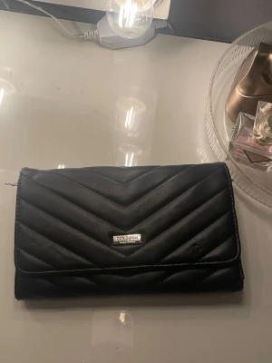 Svart kuvertväska  - Snygg svart kuvertväska från Don Donna med quiltad design i chevronmönster. Väskan är tillverkad i skinnimitation och har en silverfärgad logodetalj på framsidan. Perfekt storlek för att bära med det viktigaste och passar till många olika stilar.