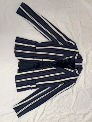 Randig kavaj från GANT, blå/vit - Snygg kavaj från GANT i blått och vitt med vertikala ränder. Klassisk krage, två knappar framtill och fickdetaljer. Materialet är en mix av polyester, viskos och elastan som ger en skön och flexibel känsla. Perfekt för dig som gillar stilrena och trendiga plagg.