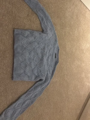 Ljusblå stickad tröja från LMTD - Säljer en ljusblå stickad tröja från LMTD i storlek S. Tröjan har ett snyggt hålmönster med diamantformade detaljer och ribbade muddar vid hals, ärmslut och nederkant. Perfekt för dig som gillar mjuka och mysiga plagg med lite extra stil.