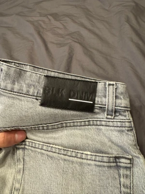 Ljusgrå jeans från BLK DNM - Säljer ett par ljusgrå jeans från BLK DNM med klassisk femficksdesign och raka ben. Jeansen har en stilren look med svart patch i midjan och är tillverkade i slitstarkt bomullsmaterial. Perfekt för dig som gillar enkel och clean stil. Litet hål vid bild 2 finns.
