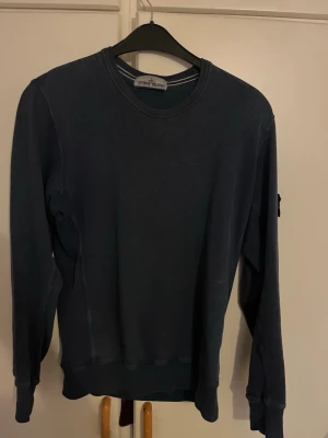 Mörkblå sweatshirt från Stone Island - Säljer en mörkblå långärmad sweatshirt från Stone Island med klassisk rund hals och ikonisk patch på ärmen. Tröjan är i mjuk bomull och har en clean, loose passform som är perfekt för chill dagar. Märkets logga sitter fast med knappar på vänster ärm.