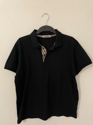 Svart Burberry piké med rutig detalj - Säljer en svart pikétröja från Burberry med klassisk krage och korta ärmar. Tröjan har en snygg rutig detalj vid knäppningen i typisk Burberry-stil. Perfekt för dig som gillar stilrena och tidlösa plagg med en twist.