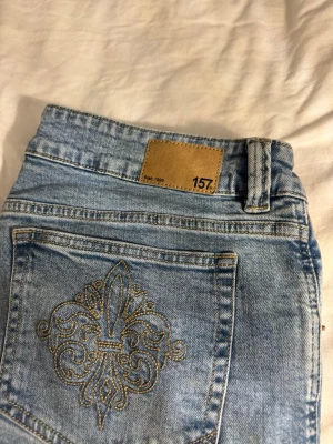 Ljusblå jeans med broderad ficka från 157 - Snygga ljusblå jeans från 157 med rak passform och klassisk femficksdesign. Bakfickan har ett detaljerat broderi i guldtråd som ger extra stilpoäng. Jeansen har knapp och dragkedja framtill samt bälteshällor. Perfekta för dig som gillar en chill och trendig look. Och de är låg midjade