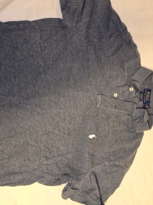 Grå pikétröja från Polo Ralph Lauren - Snygg grå pikétröja från Polo Ralph Lauren med klassisk krage och två vita knappar. Tröjan har korta ärmar och den ikoniska broderade loggan på bröstet. Perfekt för en chill och stilren look.