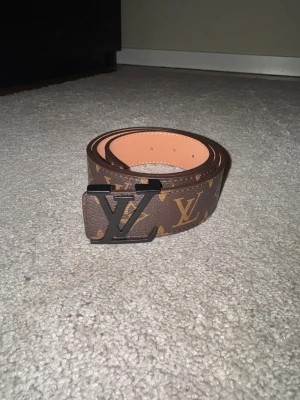 Lyxigt brunt monogrammönstrat bälte med svart spänne - Snyggt och exklusivt bälte i klassiskt brunt monogrammönster med ett stilrent svart metallspänne som ger en modern och självsäker look. En riktig statement-accessoar som lyfter vilken outfit som helst – lika snygg till jeans som till mer dressade plagg.  Bältet har en tidlös design, känns gediget och är i fint skick. Perfekt för dig som vill addera en lyxig touch till vardagen utan att kompromissa med stil.