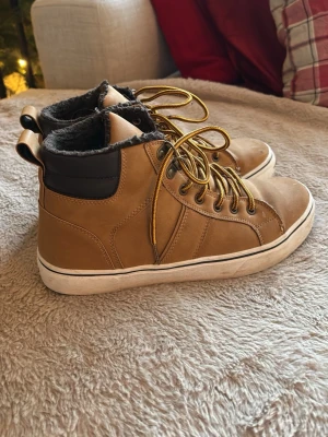 Bruna sneakers från Everest med foder - Säljer ett par bruna sneakers från Everest med varm, mjuk fuskpäls på insidan. Skorna har gulbruna snören, svart detalj vid ankeln och platt vit sula. Utsidan är i skinnimitation och de har rund tå. Perfekta för kyliga dagar.