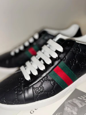 Svarta sneakers från Gucci med GG-mönster - Säljer ett par svarta Gucci sneakers i skinn med präglat GG-mönster. Skorna har vita snören, vit sula och klassiska gröna och röda ränder på sidan. Gucci-logga i silver på hälen och detaljerad sula. Perfekt för dig som gillar lyxiga och snygga sneakers.
