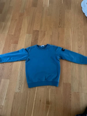 Blå sweatshirt från Stone Island Junior - Säljer en blå sweatshirt från Stone Island Junior med klassisk rund hals och långärmad passform. Tröjan har den ikoniska Stone Island-patchen på vänster ärm och är tillverkad i mjuk bomull. Perfekt för dig som gillar streetwear och snygga detaljer. Har dock blivit tvättad med märket på som gör det lite blå aktigare.