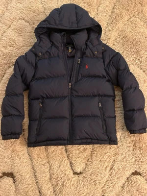 Mörkblå pufferjacka från Polo Ralph Lauren - Säljer en mörkblå pufferjacka från Polo Ralph Lauren i storlek S. Jackan har huva, dragkedja framtill och två fickor med dragkedja. Klassisk röd Polo-logga på bröstet och quiltad design som håller värmen. Perfekt för kalla dagar!