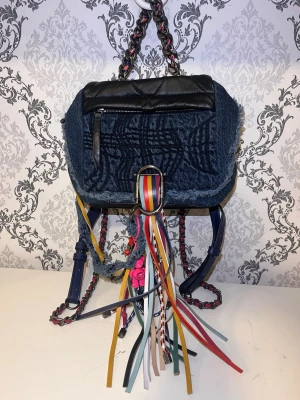 Blå ryggsäck med fransar från Diesel - Unik ryggsäck från Diesel i blått denim och svart skinn med coola fransdetaljer i flera färger. Väskan har kedjehandtag med inslag av rosa och blått, dragkedja fram och råa kanter. Insidan är svart med innerfack och Diesel-logga. Tyckte inte att den var fin, väskan är oanvänd.