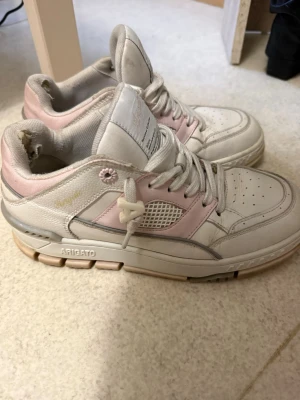 Axel Arigato sneakers i vitt och rosa - Säljer ett par snygga Axel Arigato sneakers med vit bas och ljusrosa detaljer. Skorna har snörning, rund tå och meshpaneler på sidorna. Sulan är chunky och beige, och det finns en liten blomdetalj vid snörningen. Materialet är skinn och textil. Säljer dessa då de är för stora för mig.