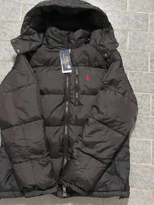 Svart pufferjacka från Polo Ralph Lauren - Svart pufferjacka från Polo Ralph Lauren med huva, två sidofickor med dragkedja och klassisk röd logga på bröstet. Jackan har quiltad design och är tillverkad i polyester, perfekt för kalla dagar. Snygg och stilren look med normal passform.