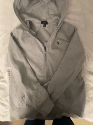 Grå Polo Ralph Lauren zip hodde - En stilren grå Polo Ralph Lauren zip hoddie. Väldigt bra skick utan bara en liten defekt längst ner vid tröjan där det finns ett litet blåt märke. Den är Xl i barn storlek och passar mig ganska bra då jag är 172. Hade sagt det passar en från 170 ner till 160. Om du har några frågor eller liknade så hör gärna av dig🙌