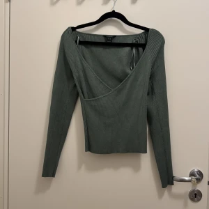 Grön omlottopp från Lindex - Snygg ribbad grön omlottopp från Lindex i storlek M. Toppen har lång ärm, djup v-ringning och knytband vid axeln för extra detalj. Perfekt att styla med jeans eller kjol för en trendig look. Säljer likadan i vitt. 