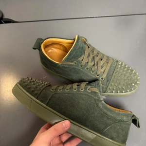 Gröna Christian Louboutin sneakers med nitar - Unika gröna sneakers från Christian Louboutin i mocka med coola nitar på tån och klassisk röd sula. Skorna har snörning och en rund tå, samt beige insida i skinn. Perfekt för dig som vill sticka ut med exklusiv stil och edgy detaljer.