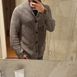 Gråmelerad kofta från - Säljer en gråmelerad stickad kofta från H&M L.O.G.G. med stora knappar och två fickor framtill. Koftan har en bred krage och är långärmad, perfekt för lager-på-lager under kyliga dagar.