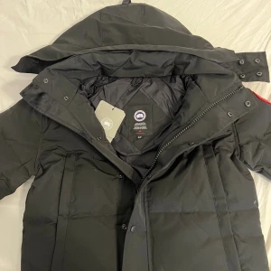 Svart dunjacka från Canada Goose - Svart dunjacka från Canada Goose med stor huva och klassisk logga på ärmen. Jackan har dragkedja och knappar framtill, samt quiltad design för extra värme. Perfekt för kalla dagar och har en clean, stilren look som funkar till allt.