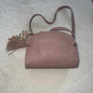 Rosa axelväska från Forever New - Söt rosa axelväska från Forever New i syntetmaterial med justerbar rem och dekorativ tofs i sidan. Väskan har guldfärgade metalldetaljer och dragkedja upptill. Perfekt storlek för det viktigaste och stilren design.